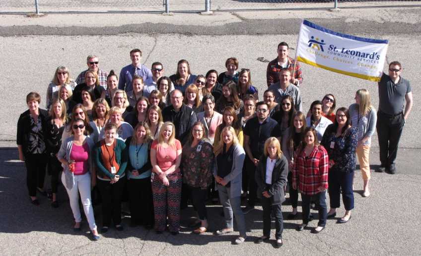 The St. Leonard's Community Services staff outside in a group holding an agency branded flag. 